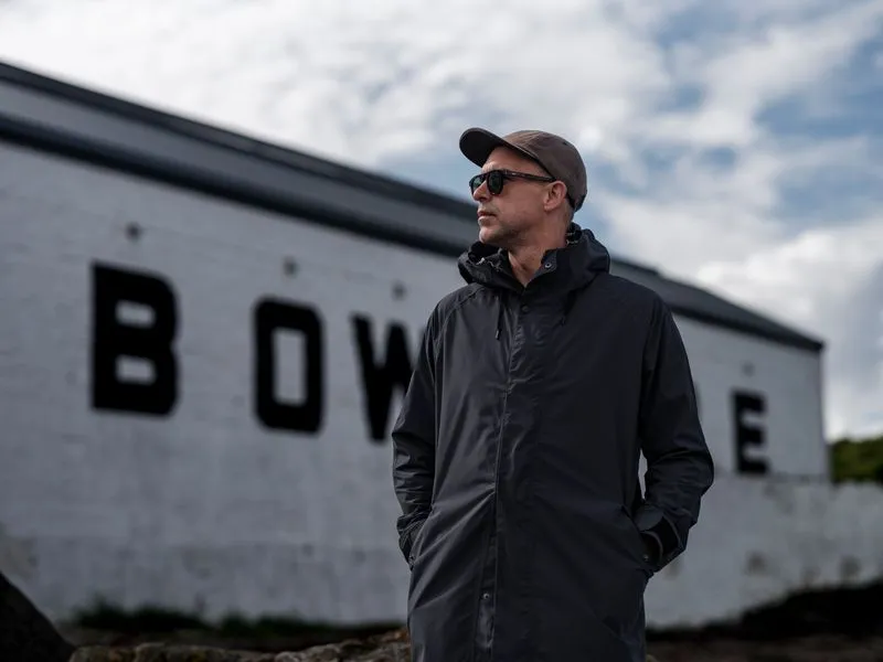 A person wearing a dark waterproof jacket, baseball cap, and sunglasses stands with their hands in their pockets outside a white industrial building. Large black letters spelling part of "BOWMORE" are visible on the building behind them. The person is looking off into the distance under a cloudy sky, creating a calm, contemplative atmosphere.