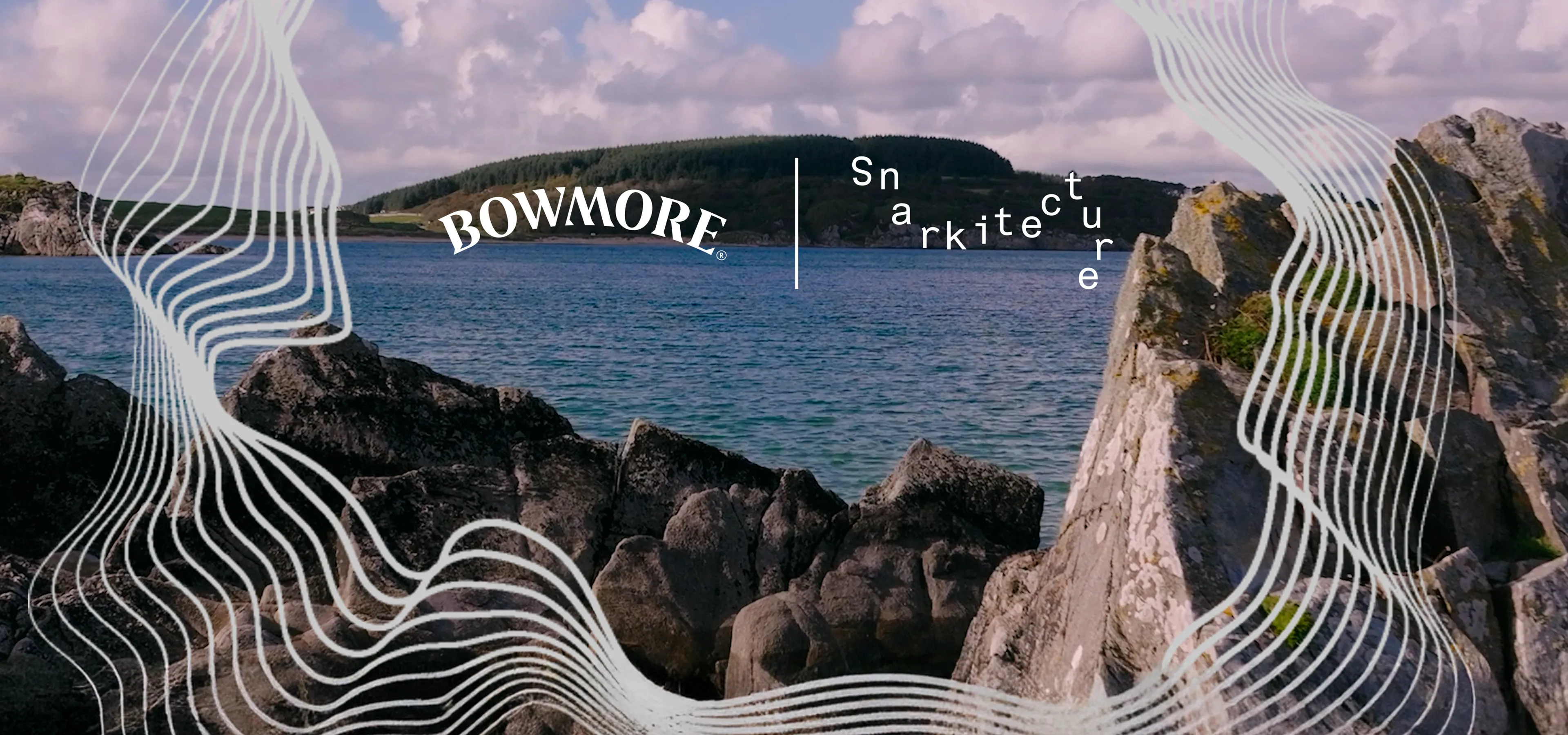 A rocky shoreline overlooks a calm bay with a tree-covered island in the distance beneath a blue sky with soft clouds. White flowing contour lines frame the top and bottom of the image, while the Bowmore and Snarkitecture logos appear near the center, highlighting the collaboration against the coastal Islay landscape.